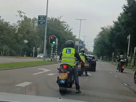 Leerlingen met een geel hesje en een L-bord rijden bij groen licht weg op een stadsweg in Utrecht, gezien vanuit de auto erachter.